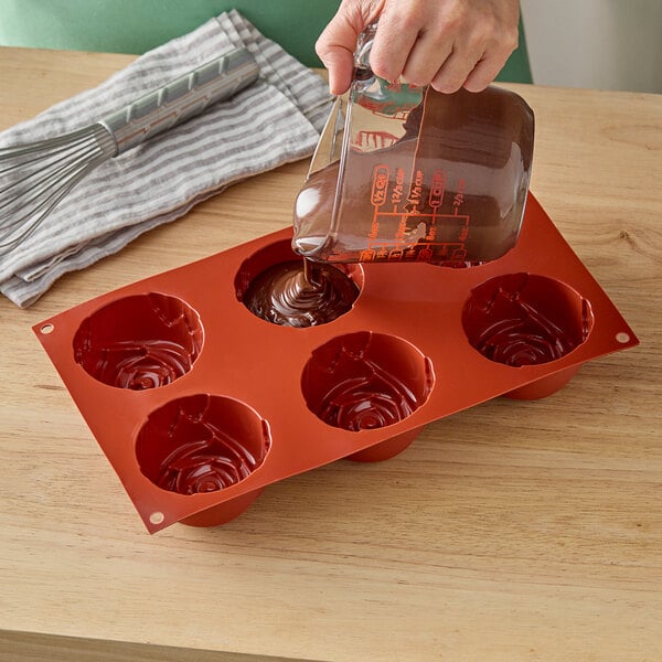 A hand pouring chocolate into a Silikomart rose silicone baking mold.