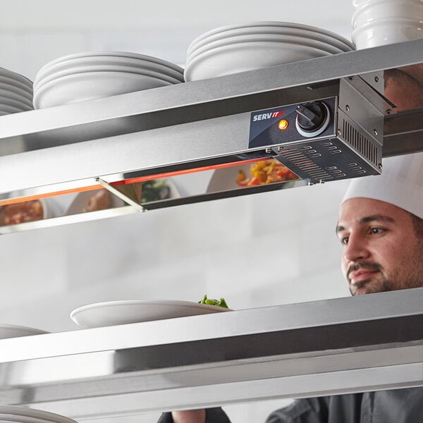 A chef in a professional kitchen looking at a shelf of white plates above a ServIt strip warmer.