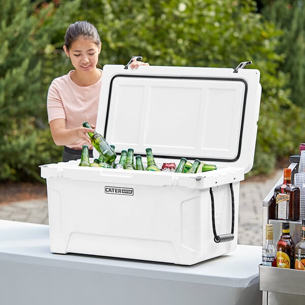 A woman opening a white CaterGator outdoor cooler to put green bottles inside.