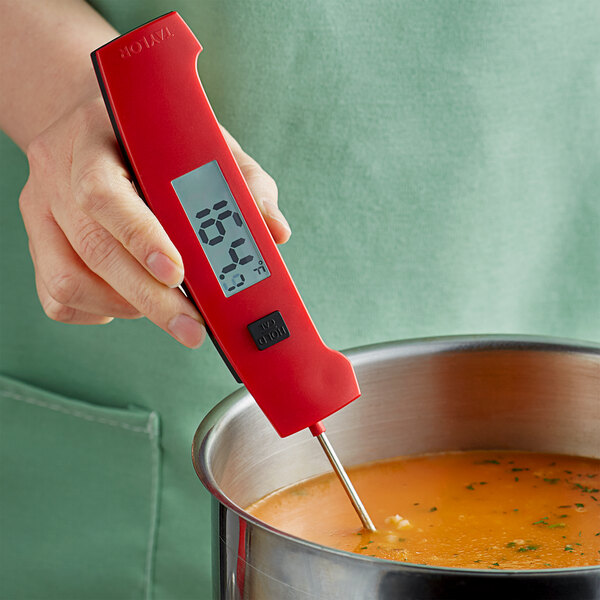 A person holding a Taylor red digital thermocouple thermometer over a pot of soup.