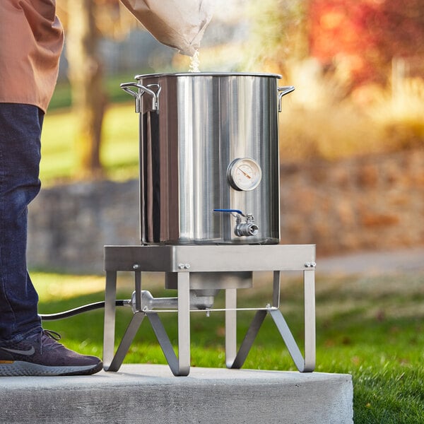 A person pouring water into a large stainless steel brewing pot on a metal structure with metal legs.