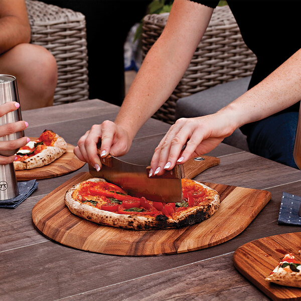 A hand using a Fox Run acacia wood pizza peel to cut a pizza on a wood surface.