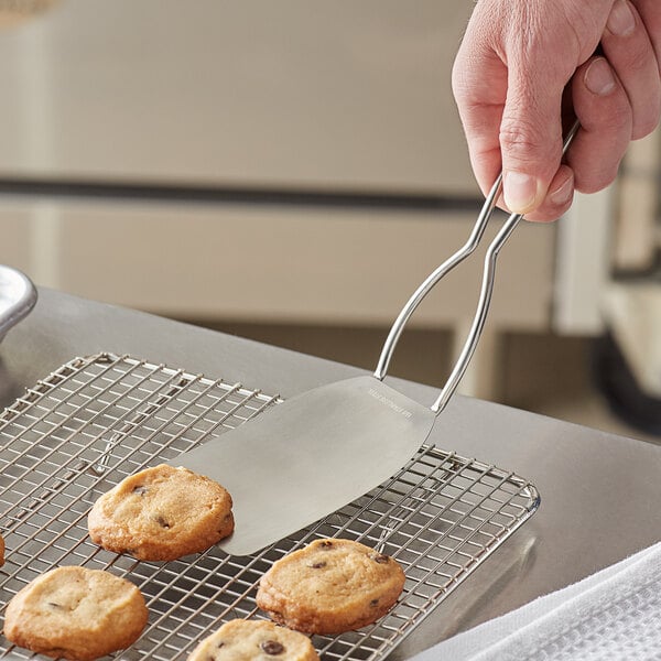A person holding an Ateco stainless steel spatula over a tray of cookies.