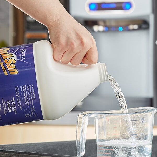 A hand pouring Noble Chemical QuikSan sanitizer into a measuring cup on a counter.
