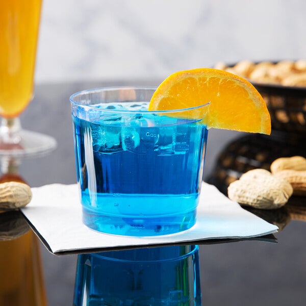 A close up of a WNA Comet Classicware clear plastic fluted tumbler filled with a blue drink and an orange slice.