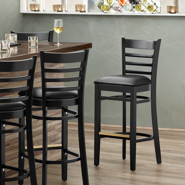 A Lancaster Table & Seating black wood ladder back bar stool with black vinyl seat.
