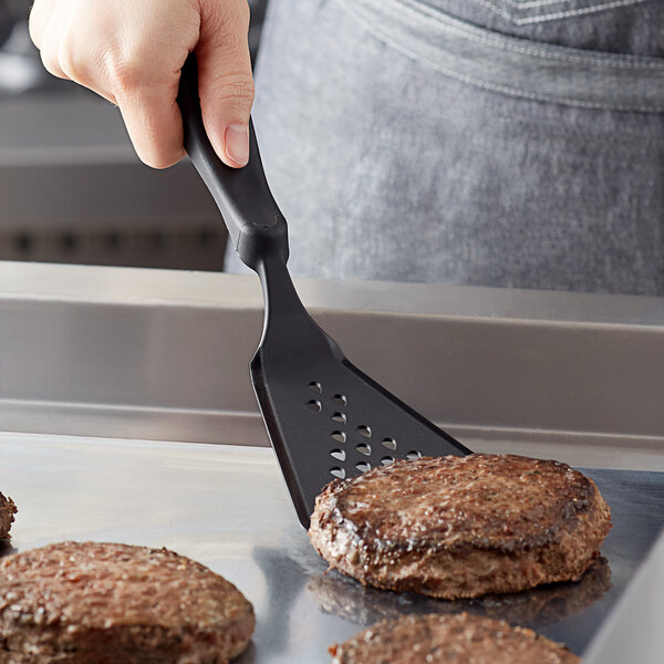 A hand using a Linden Sweden black silicone spatula to flip burgers in a pan.