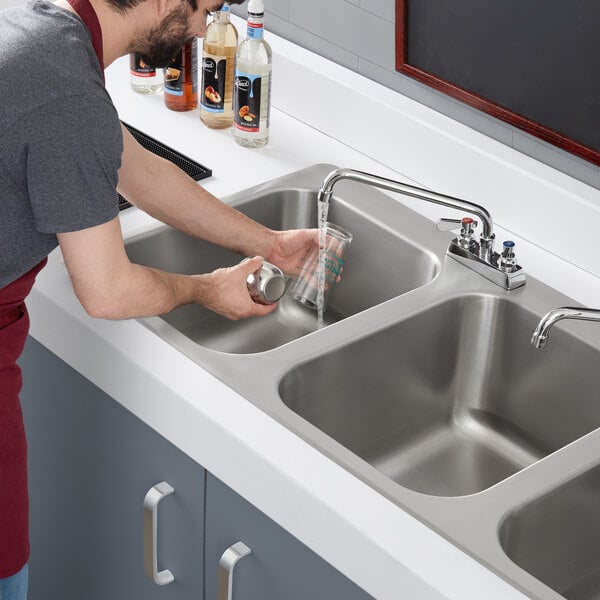 A man washing a glass in a Regency stainless steel drop-in sink with three compartments.