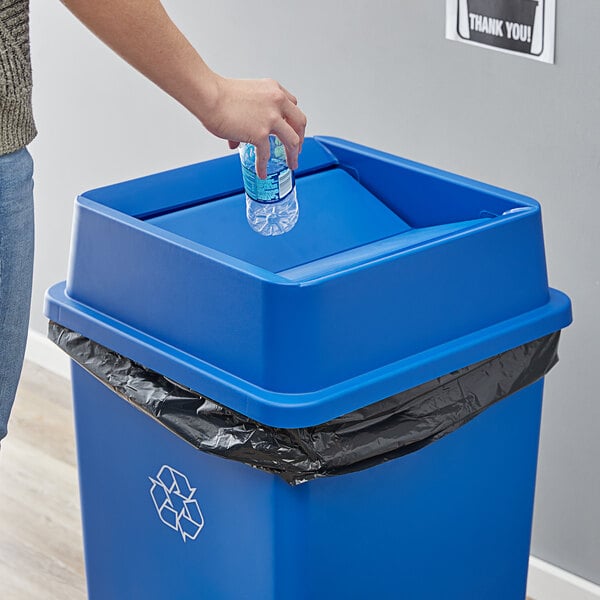A person throwing a plastic bottle into a blue Lavex trash can with a swing lid.