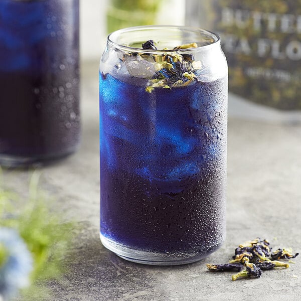 A glass of blue butterfly pea flower lemonade with ice and dried hibiscus flowers on the table.