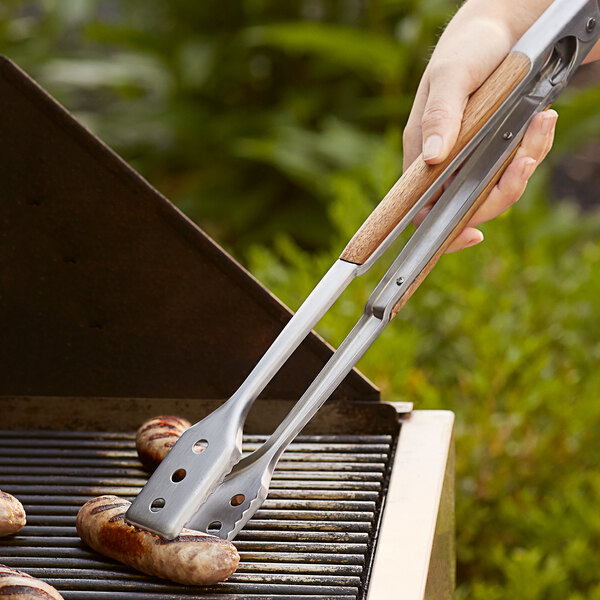 Outset stainless steel tongs with acacia wood handles being used to cook sausages on a grill.