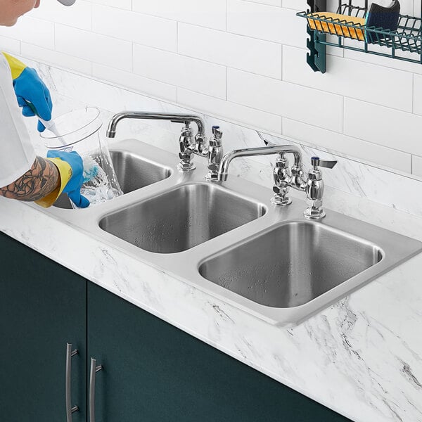A person in blue gloves washing dishes in a Waterloo stainless steel drop-in sink.