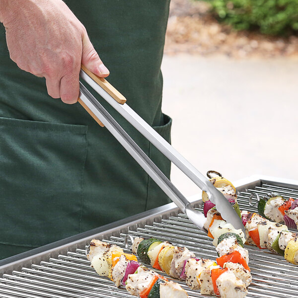A person using Outset stainless steel tongs to hold skewers over a grill.