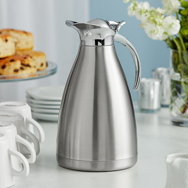 An Acopa stainless steel coffee carafe on a table with white coffee mugs.