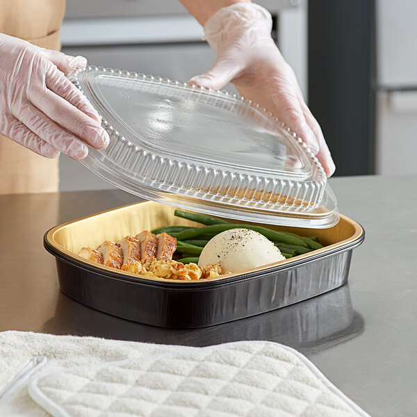 A person in gloves placing a plastic lid on food in a black and gold aluminum foil container.