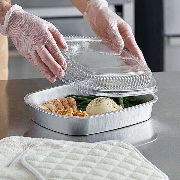 A person in gloves holding a ChoiceHD Smoothwall silver foil entree pan with a dome lid over food.