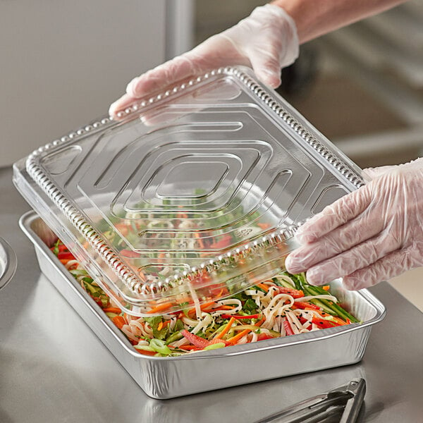 A person holding a ChoiceHD silver foil tray with a plastic dome lid over food.