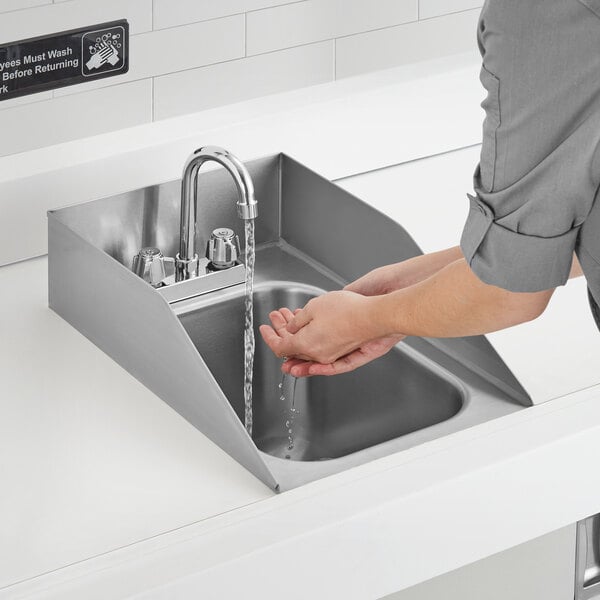 A person washing their hands in a Regency stainless steel sink with gooseneck faucet and side splashes.