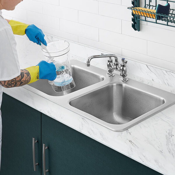 A person in yellow and blue gloves cleaning a Waterloo stainless steel two compartment sink.