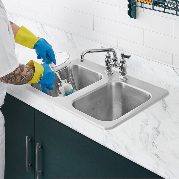 A person wearing blue rubber gloves washing a glass in a Waterloo stainless steel two compartment sink.