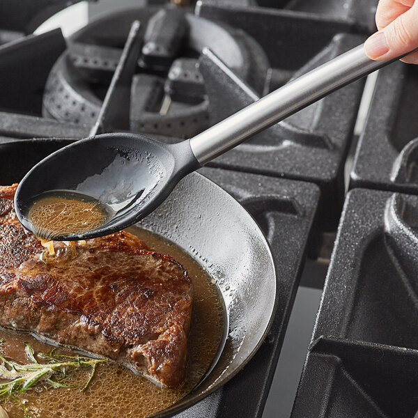 A Tablecraft stainless steel spoon with black silicone head pouring sauce on a meat dish.