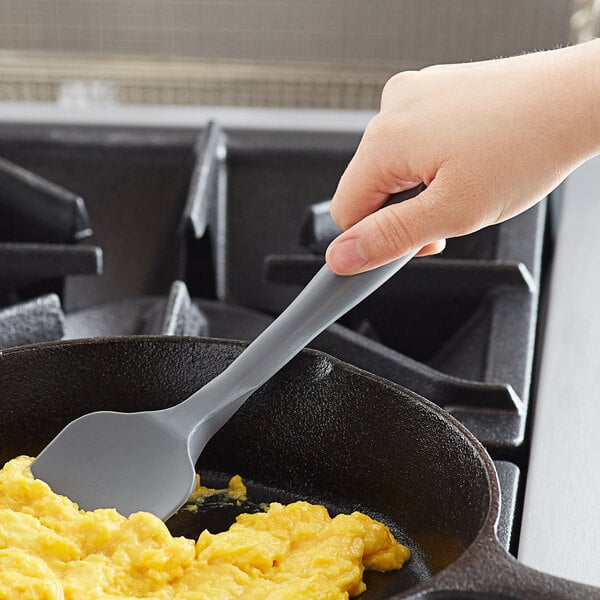 A hand using a Tablecraft gray silicone spoonula to stir scrambled eggs in a pan.