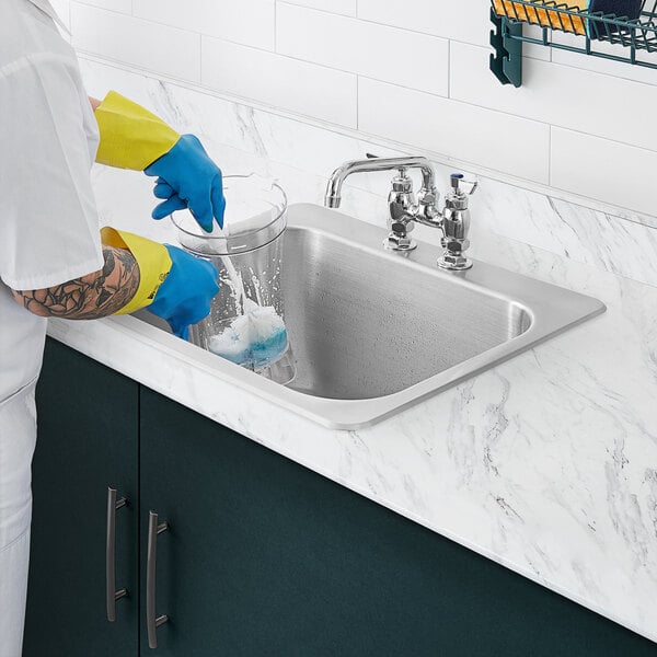 A person wearing blue gloves uses a toothbrush to clean a Waterloo stainless steel sink.