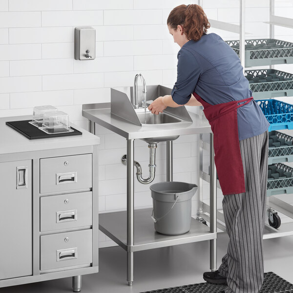 A woman in a red apron washing a Regency stainless steel sink in a school kitchen.
