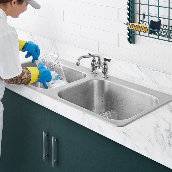 A person wearing blue gloves washing dishes in a Waterloo stainless steel two compartment sink.