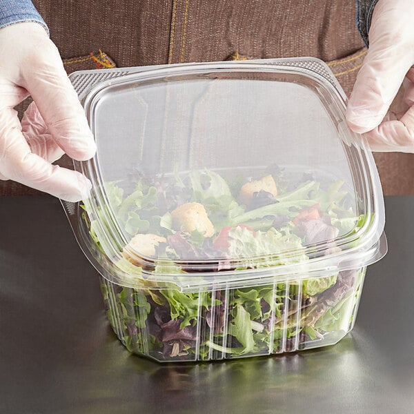 A person in gloves holding a Choice clear plastic deli container with a salad inside.