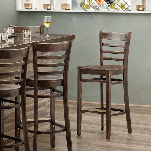 A Lancaster Table & Seating wood ladder back bar stool with a vintage wood seat on a table with a glass of wine.