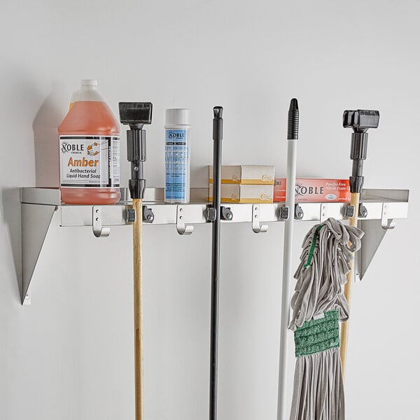 A Regency stainless steel utility shelf with mop and broom holders and rag hooks holding cleaning supplies and tools.