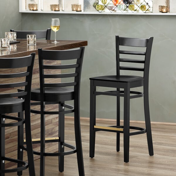 A Lancaster Table & Seating black wood ladder back bar stool with a black wood seat next to a table with glasses on it.