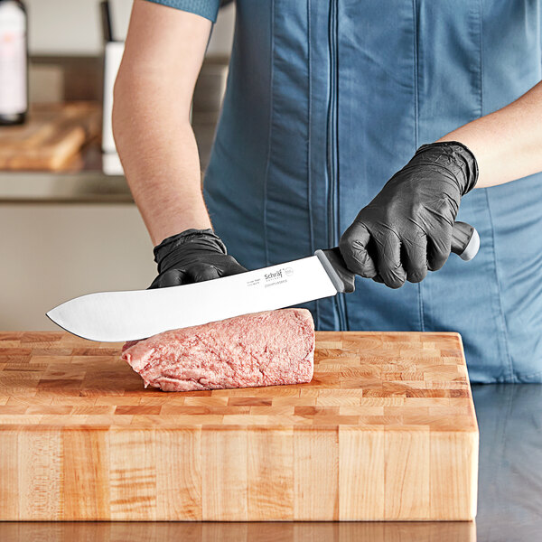 A person in gloves using a Schraf butcher knife to cut meat on a cutting board.