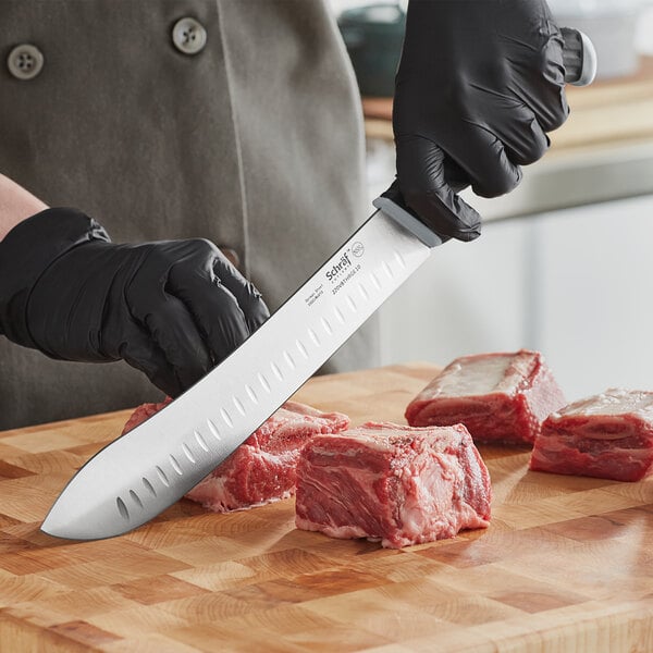 A person wearing black gloves uses a Schraf butcher knife to cut meat on a cutting board.