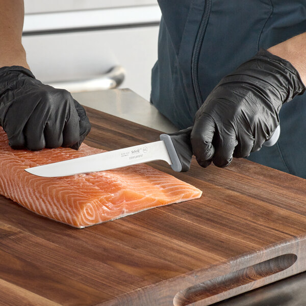 A person wearing black gloves uses a Schraf semi-flexible fillet knife to cut a piece of salmon.