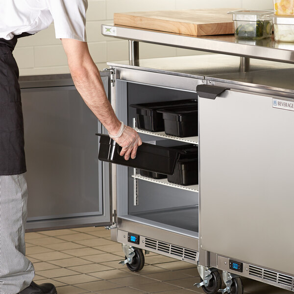 A man in a glove opening the left-hinged door of a Beverage-Air undercounter freezer to put a black tray inside.