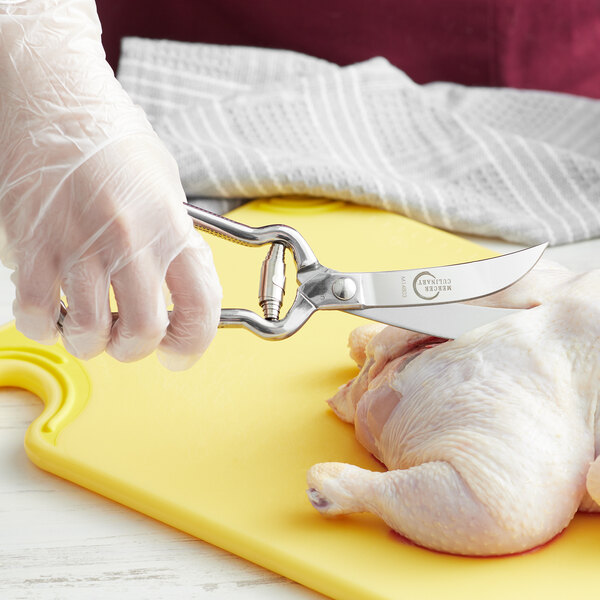A hand using Mercer Culinary stainless steel poultry shears to cut a chicken on a yellow cutting board.