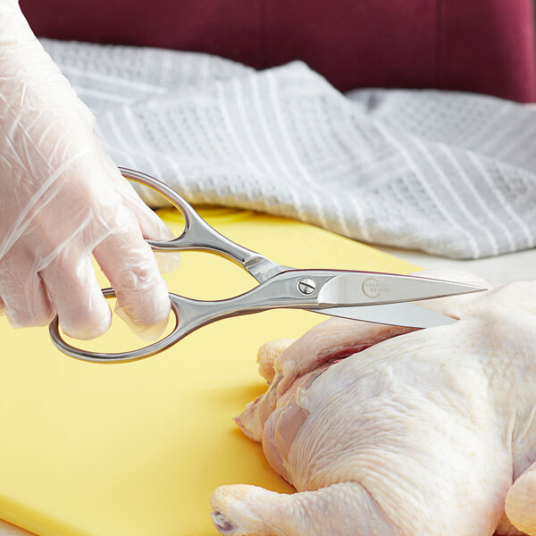 A gloved hand using Mercer Culinary stainless steel kitchen shears to cut a chicken.