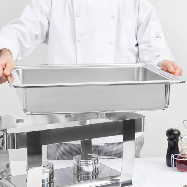 A chef holding a Vollrath stainless steel water pan on a tray above a glass of liquid.
