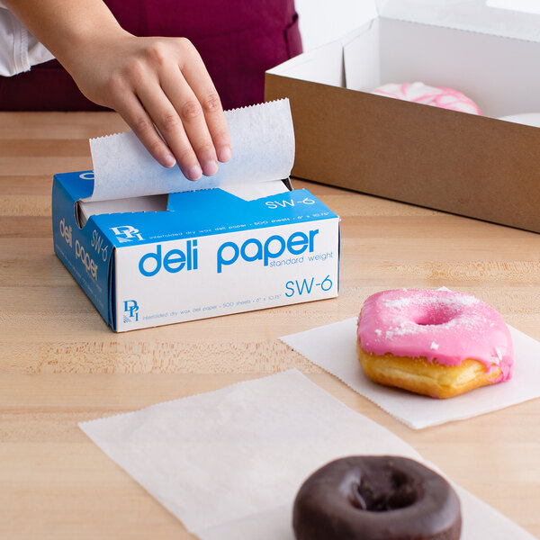 A person using Durable Packaging wax paper to pick up a pink donut with sprinkles.