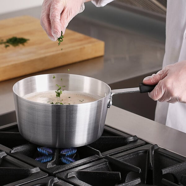 A hand stirring food in a Choice aluminum sauce pan on a stove.