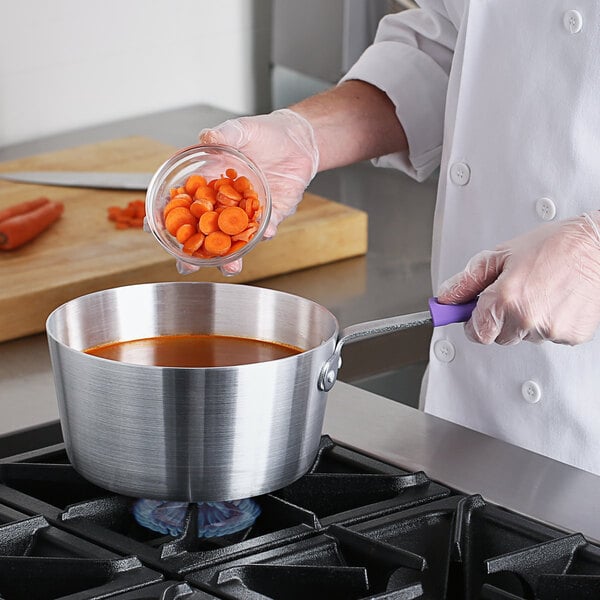 A person in a white coat pouring sliced carrots into a Choice aluminum sauce pan on a stove.
