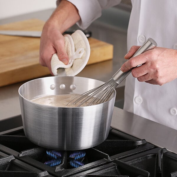 A chef using a whisk to mix food in a Choice aluminum sauce pan on a stove.