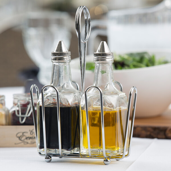 A Tablecraft clear glass oil and vinegar cruet set in a chrome rack on a table with oil and vinegar bottles inside.
