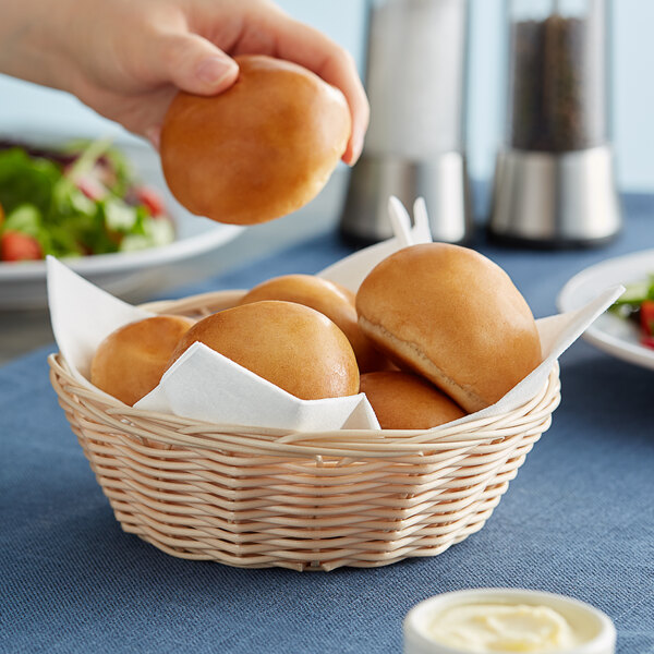 A hand holding a bun from a Tablecraft natural-colored polypropylene bread basket.