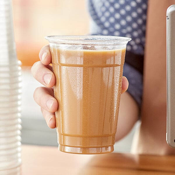 A person holding a Choice Heavy Weight clear plastic cup with a straw filled with a brown liquid.