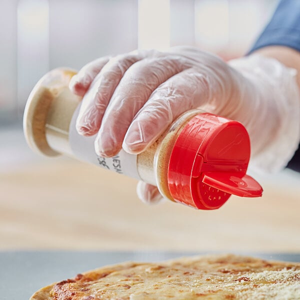 A gloved hand pouring pepper from a round plastic spice container with a red lid with 13 holes onto a pizza.