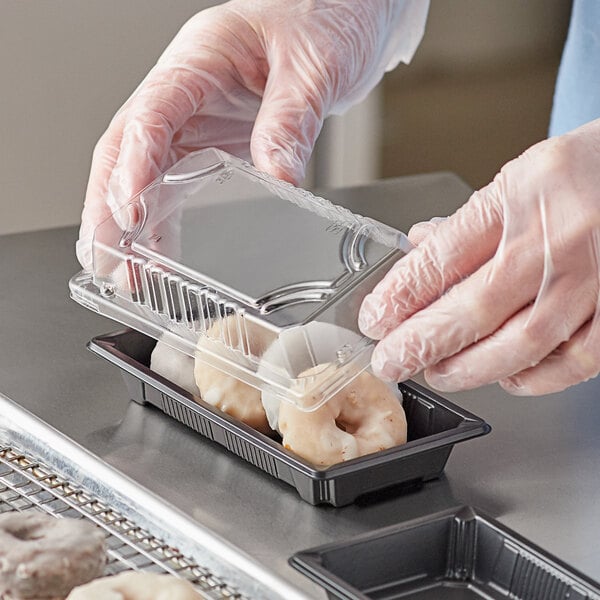 A person in gloves holding a Choice small rectangular plastic food container filled with doughnuts.