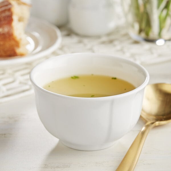 A white Acopa porcelain bouillon cup filled with soup and a spoon on a table with bread.